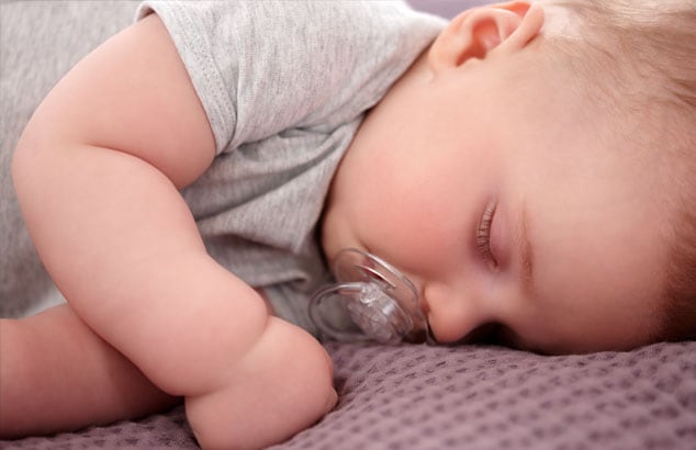 A baby with a clear, unpatterned dummy in their mouth sleeps on their side on a purple blanket.