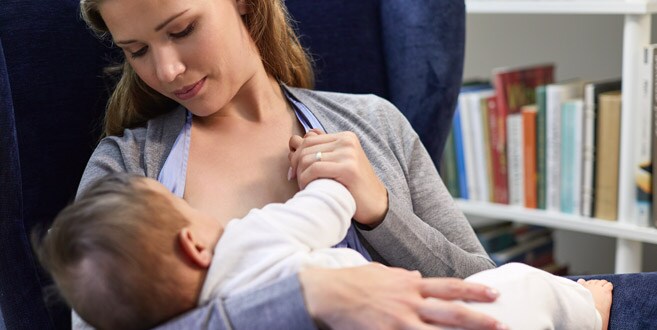 A mother is sat back in a blue velvet chair looking down at her breastfeeding baby demonstrating the cradle hold.