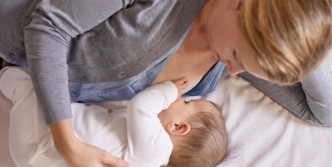 A mother is lying on her left side on a white bed, cradling a baby breastfeeding in lying down position.