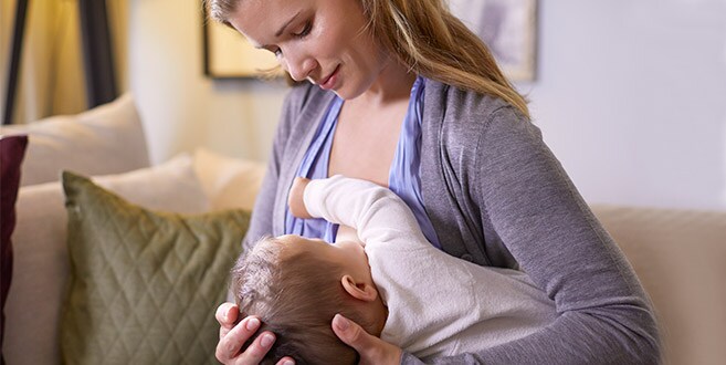 A mother is sat upright on a cream sofa, looking down at her breastfeeding her baby using the underarm hold.