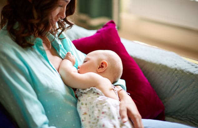 A mother wearing a blue shirt breastfeeds her baby in a seated position on a comfortable grey sofa.