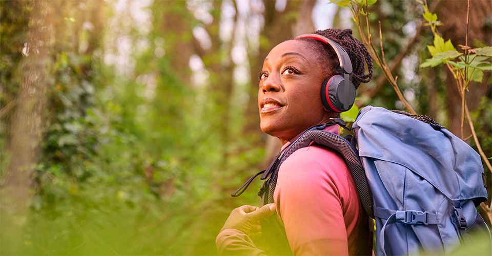Woman hiking through a forest with a blue backpack, wearing Philips A6219 headphones and looking up with a smile