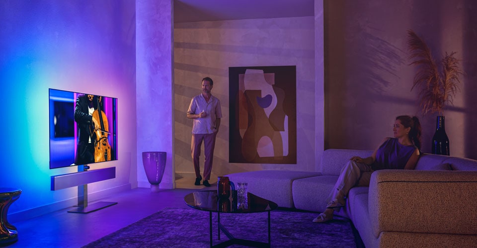 Man and woman in a stylish living room watching a Philips Ambilight TV displaying a cello performance
