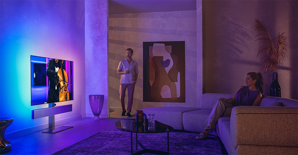 Man and woman in a stylish living room watching a Philips Ambilight TV displaying a cello performance
