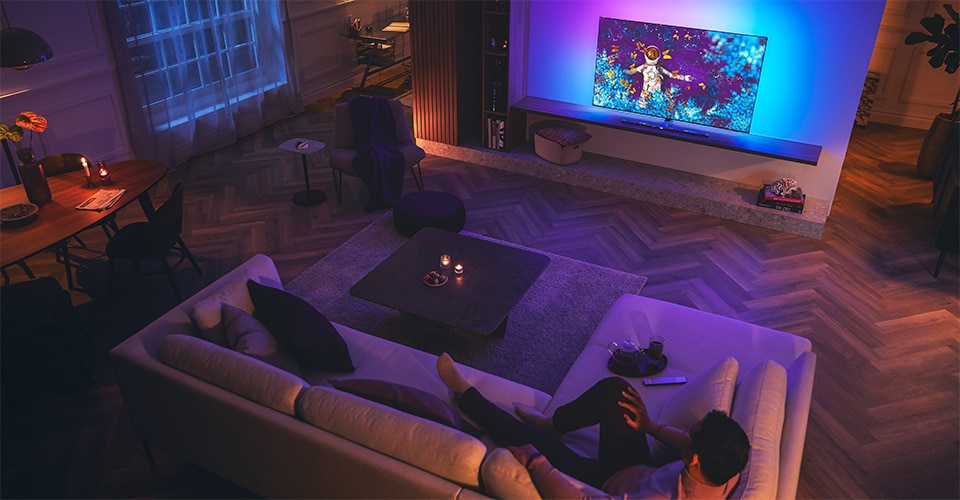 Man relaxing on a sofa in a dimly lit living room, watching a Philips Ambilight TV