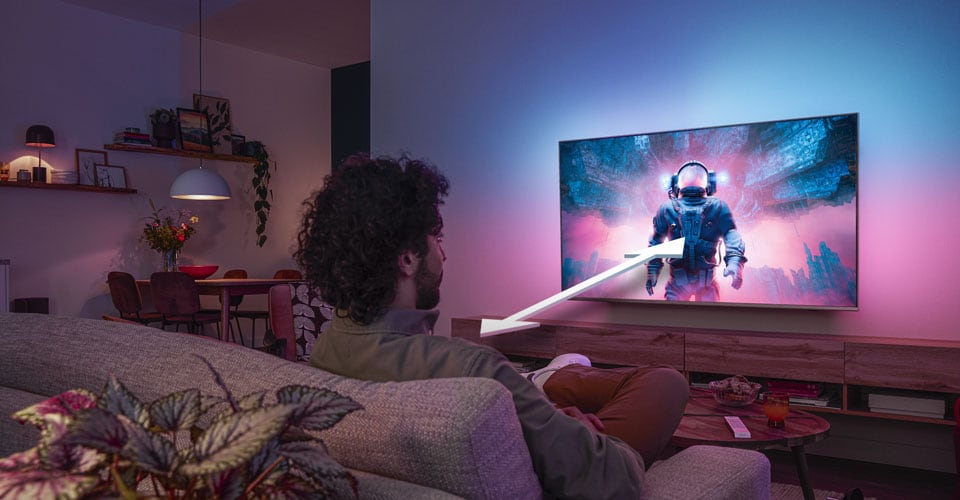 Man sitting on a sofa watching a Philips Ambilight TV displaying a colorful astronaut scene, with an arrow indicating optimal viewing distance