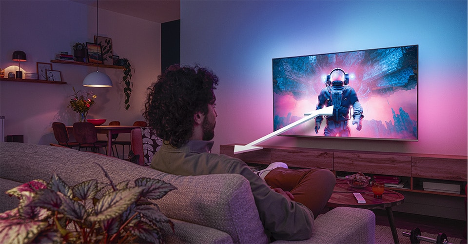 Man sitting on a sofa watching a Philips Ambilight TV displaying a colorful astronaut scene, with an arrow indicating optimal viewing distance