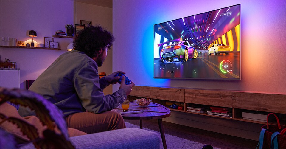 A person is sitting on a couch, playing a racing video game on a Philips Ambilight TV