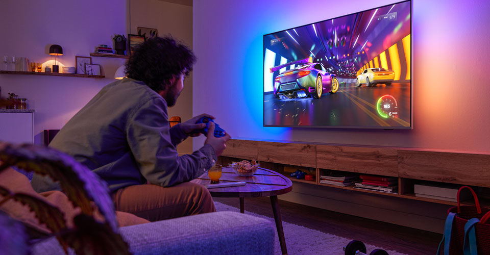 A person is sitting on a couch, playing a racing video game on a Philips Ambilight TV