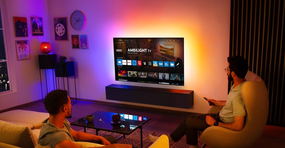 Two men watch a wall-mounted Ambilight TV in a living room
