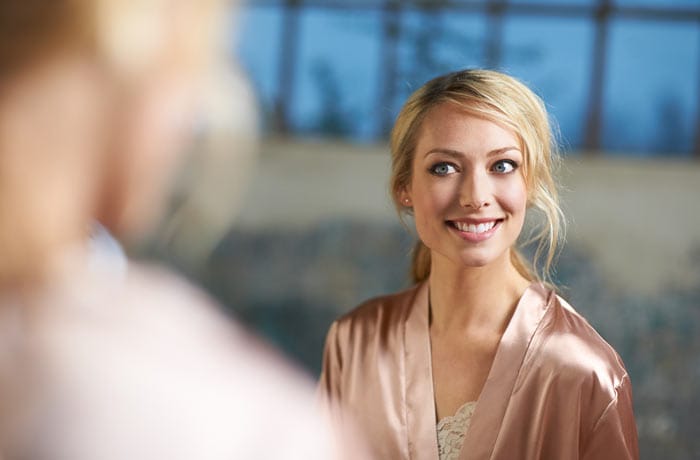 A young blonde woman looking in the mirror and looking at her white teeth.