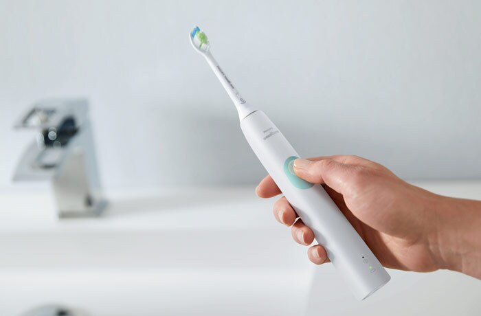 A hand holding a white Philips Sonicare electric toothbrush in front of a sink with the thumb on the button.