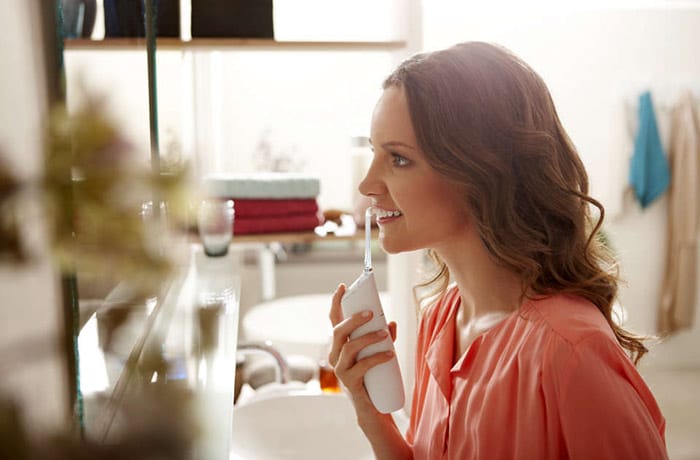 A young woman is looking into a bathroom mirror while flossing her teeth with the Sonicare Power Flosser.