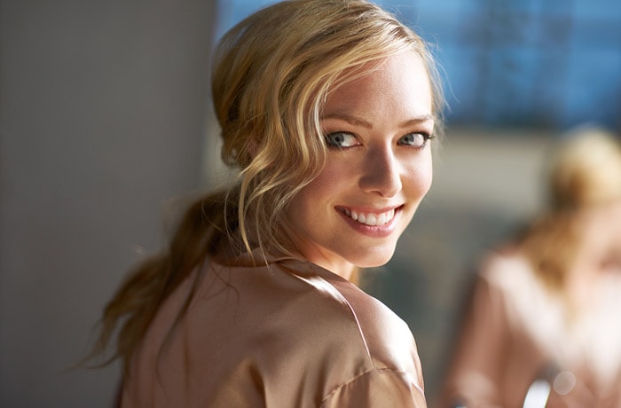 A young woman is turning toward the camera looking to the side with a smile on her face, revealing bright white teeth.
