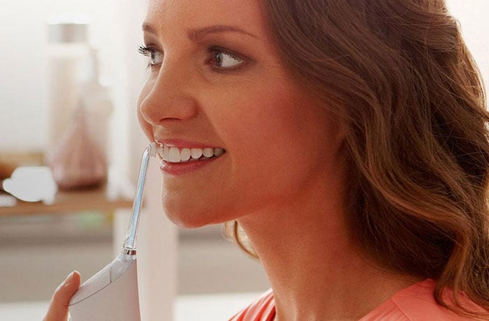 A woman is looking away from the camera brushing her white teeth with a Philips Sonicare Airfloss electric toothbrush.