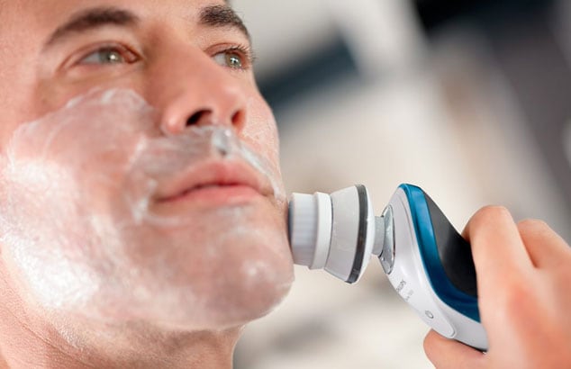 Close up of a man’s face with shaving foam on his face using an electric brush against his cheek.