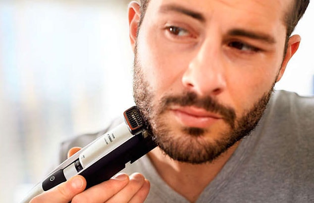 Close up of a dark-haired bearded man in grey t-shirt using an electric shaver on his jawline.