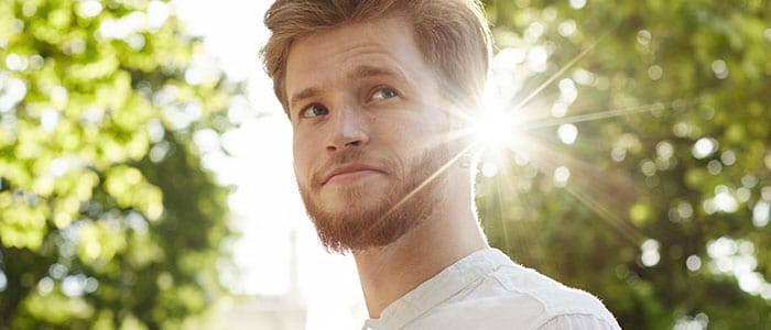 A man with light hair and facial hair only on his chin in front of sunshine and green trees.