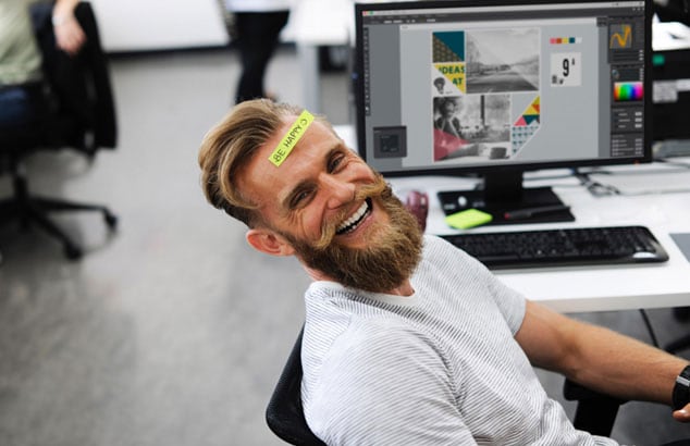 Blond, young and fun looking guy with a big beard sitting on an office chair smiling at the camera