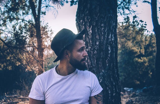 Moody image of a man in a white t-shirt and a black hat in front of a tree looking to his left.