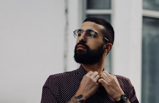 Man with a dark, full beard, nose ring, and glasses buttoning up his polka dotted dark red shirt.