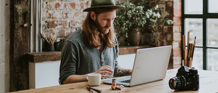 Young man with long hair and a full beard sitting in a hip space looking at a laptop in front of him.