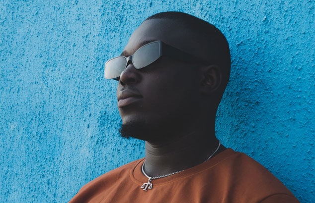 Man with a shaved head and goatee stands against a baby blue wall, wearing sunglasses.