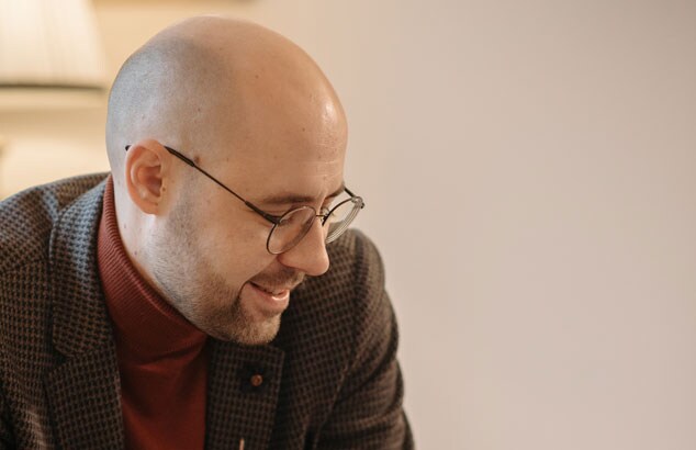 Bald man with stubble beard and moustache wears a tweed suit and glasses, while smiling and looking down.