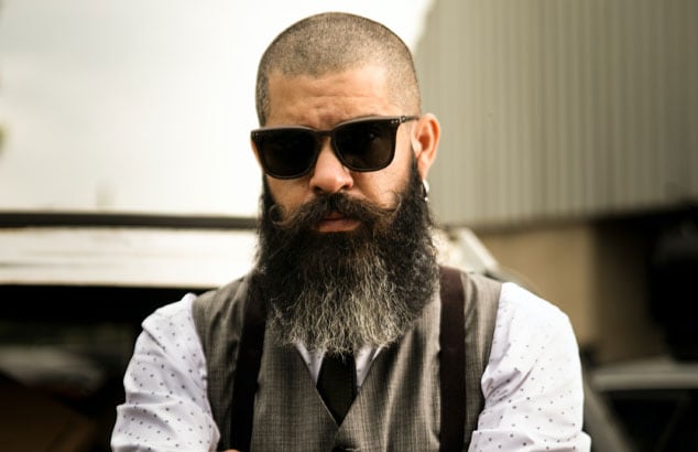 Hipster man with shaved head, long beard and handlebar moustache folds his tattooed arms while looking ahead.