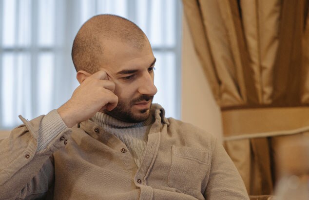 Bald man in a beige jumper and overshirt rests his stubble-covered face on his hand while sitting on a sofa.