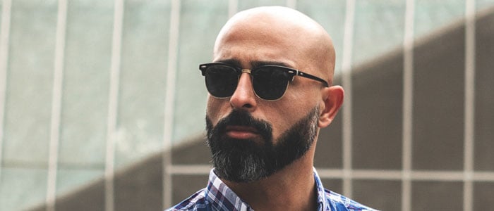 Bald man in checked shirt and sunglasses sports a thick, full beard with sharp, defined cheek lines.