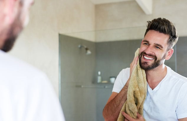 Mirror reflection of a man with dark hair and beard patting his face with a light-brown towel.