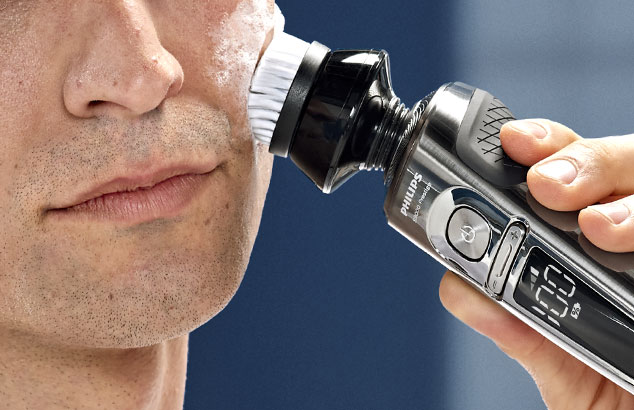 Close-up of a man with very short stubble pressing an electric brush against his cheek.