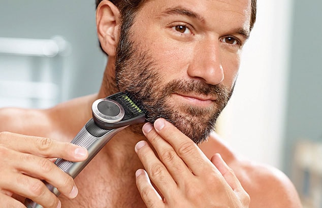 Shirtless man trimming his dark beard using a beard trimmer.