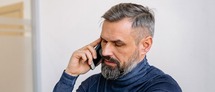 Smart, grey-haired man with a defined, medium-length pointed grey beard is on the phone while glancing at his watch.