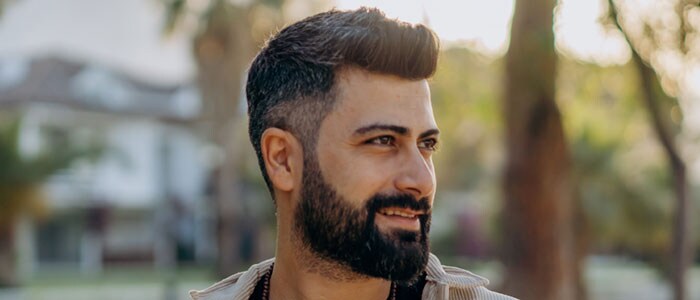 Dark-haired man with short haircut fading from sideburns to a medium length beard stands in a garden smiling.