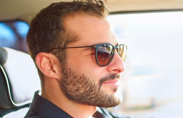 Man driving a car in black sunglasses sports a full brown beard and moustache with short haircut.