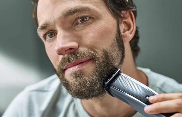 A man with a full beard shaves his face with a silver and black electric shaver.