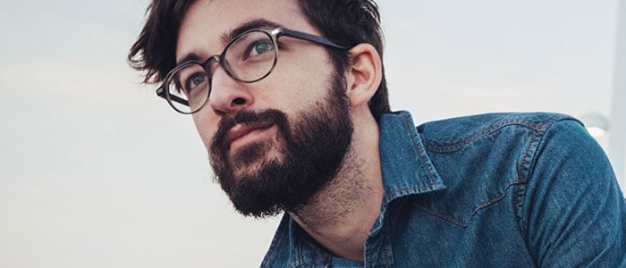Young man with a blue denim shirt, dark hair and beard and glasses is looking into the distance.