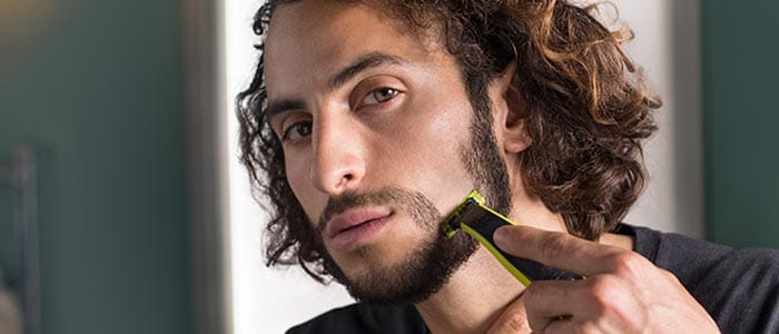 A long-haired man uses a trimmer to fully shave his beard and shape his moustache.