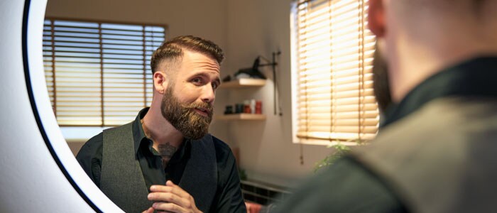 Man with a beard and a short, fade haircut smiles at himself in the mirror after cutting his own hair at home