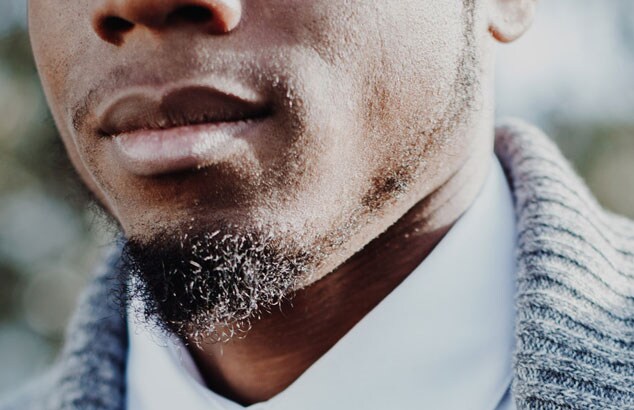 Close up of a black man in a white shirt and grey cardigan and his chin puff beard.