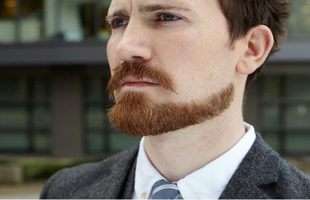 Man with an accurately cut ginger chin strap beard in a dark grey suit looks into the distance.