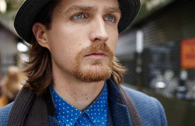 Man with shoulder long hair and henriquatre chin beard wearing a blue shirt, a suit jacket and a hat.