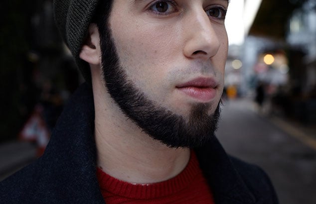 White man wearing a beanie and a red jumper with a very accurately cut dark chin curtain beard.