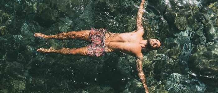 Wide shot of a bearded man in swimming shorts floating face up in water with his arms spread out.