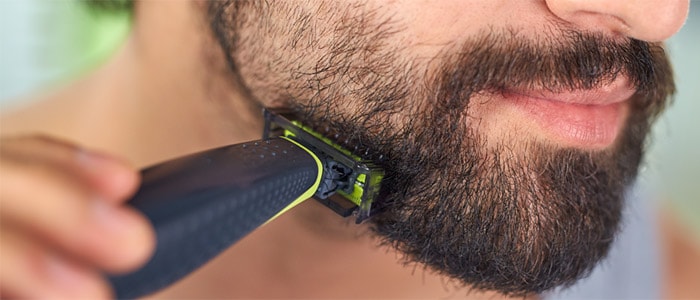 Close-up of a brown-eyed man using the Philips OneBlade to trim away the cheek hairs from his full, short beard.
