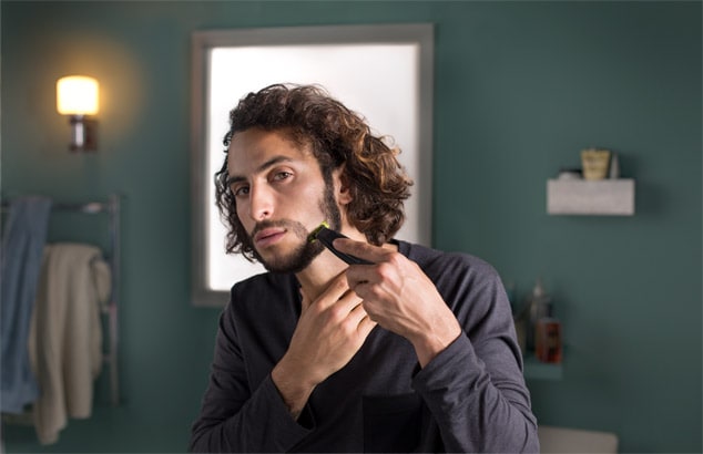 Man with shoulder-length, thick brown hair trims away cheek hair with the Philips OneBlade to shape into a goatee.