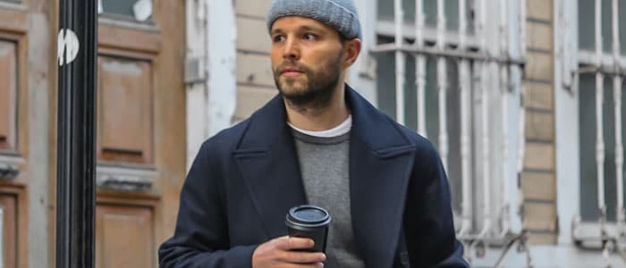 Stubble-faced man in a beanie hat and coat walks down a residential street holding a coffee cup.