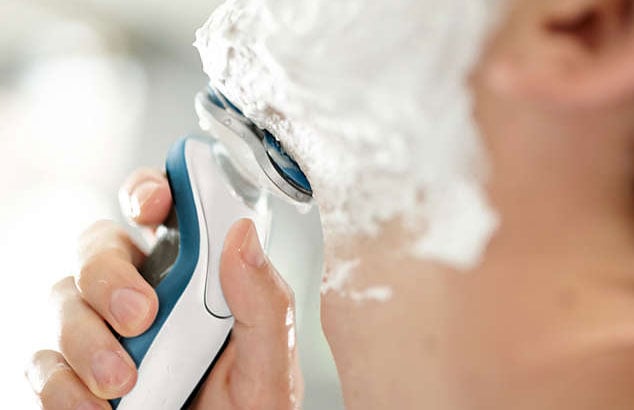 Close-up of an out-of-focus man’s chin covered with shaving foam pressing a white & blue electric shaver against his chin.
